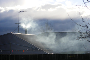 A hills hoist the foreground with the top of a dark brown fence behind it and behind that the pitched roof of a house in  with smoke billowing across it. A chimney pipe rise on the far side of the roof with smoke coming out of it. An antenna stands to the left of the chimney with dark grey clouds behind. The light of the sun is coming through the clouds at the top of the image. 