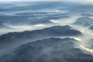 View from a plane flying over the Blue Mountains. Smoke from hazard reduction burns is visible in valleys with the dark green bush of the mountains rising above the smoke haze.