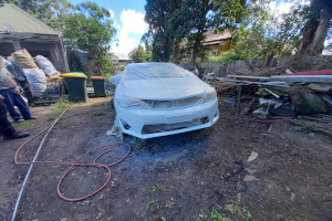 A car on a dirt area in front and to the side of a house has been sprayed with white paint. The car is facing the camera and its roof and bonnet are covered with plastic. A red hose spirals on the ground to the front and left of the car