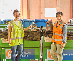 two women in hi vis vests placing old paint tins and motor oil containers in the correct recycling bins at a community recycling centre